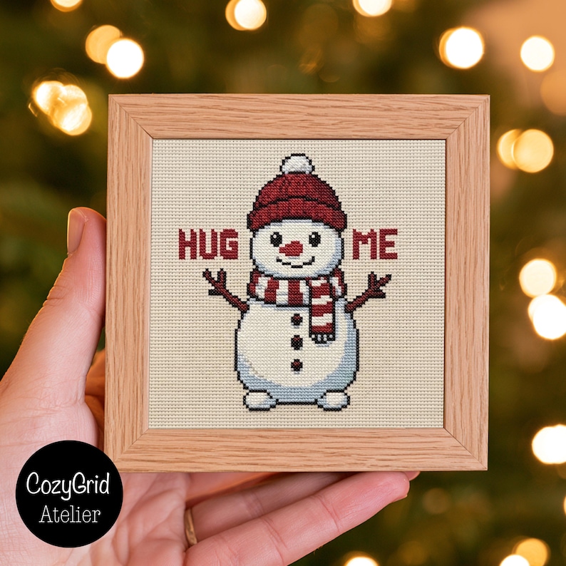 May include: A square cross-stitch embroidery in a light wood frame featuring a white snowman wearing a red knit hat and a red and white striped scarf. The snowman has stick arms, three black buttons, and the text HUG ME in red.