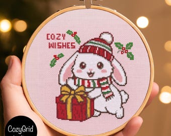 May include: A circular wooden embroidery hoop containing a cross-stitch pattern on light pink fabric. The design features a white rabbit wearing a red and green striped hat and scarf, holding a red gift with a gold bow, with the text COZY WISHES.