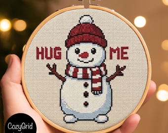 May include: A circular wooden embroidery hoop containing a cross-stitch pattern of a white snowman wearing a dark red knit hat and a matching striped scarf. The snowman has three black buttons and the text HUG ME in red block letters.