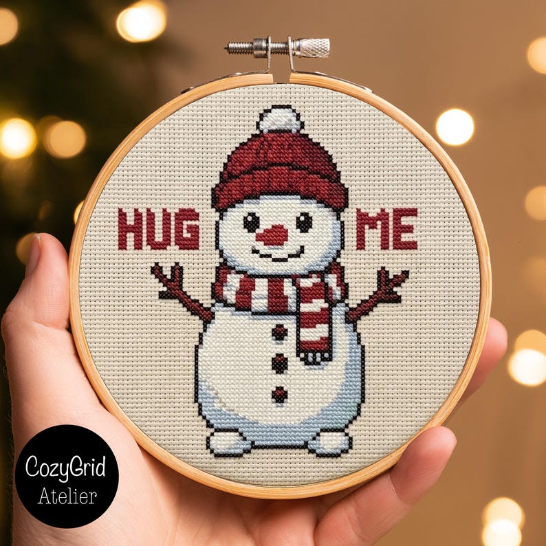 May include: A circular wooden embroidery hoop containing a cross-stitch pattern of a white snowman wearing a dark red knit hat and a matching striped scarf. The snowman has three black buttons and the text HUG ME in red block letters.