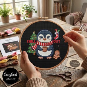 May include: A round wooden embroidery hoop containing a black fabric cross-stitch project featuring a penguin wearing a red and white striped scarf, holding a brown mug, next to a green Christmas tree, a gingerbread man, and a red stocking.