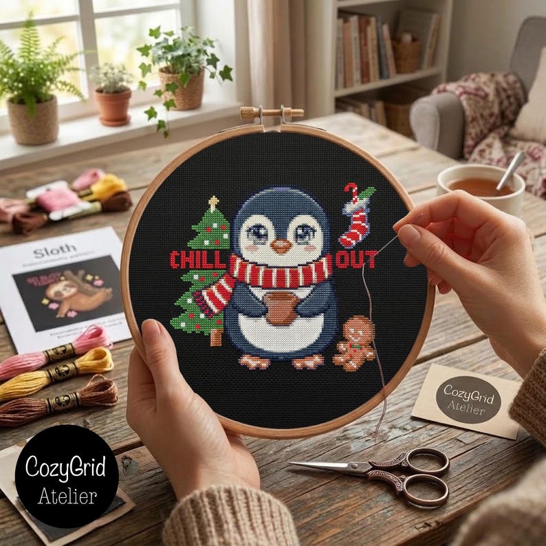 May include: A round wooden embroidery hoop containing a black fabric cross-stitch project featuring a penguin wearing a red and white striped scarf, holding a brown mug, next to a green Christmas tree, a gingerbread man, and a red stocking.