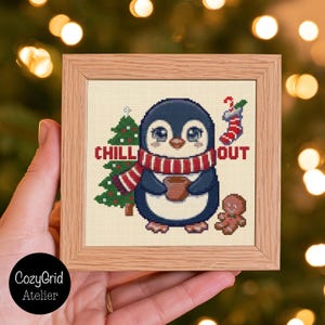 May include: A square wooden frame containing a cross-stitch pattern of a penguin wearing a red and white striped scarf, holding a brown mug. The design includes a green Christmas tree, a red and white stocking, and a gingerbread man with the text CHILL OUT.