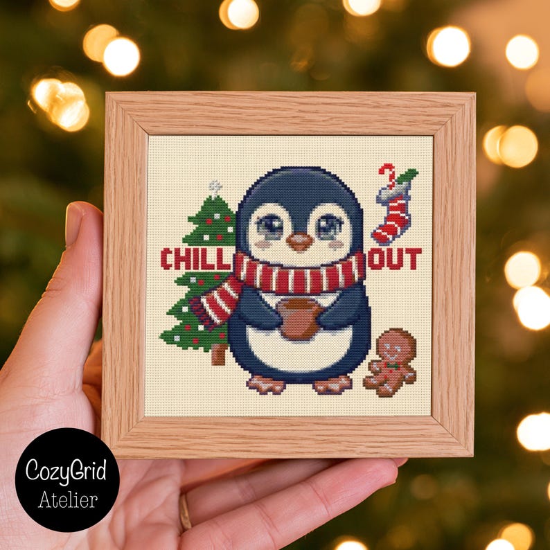 May include: A square wooden frame containing a cross-stitch pattern of a penguin wearing a red and white striped scarf, holding a brown mug. The design includes a green Christmas tree, a red and white stocking, and a gingerbread man with the text CHILL OUT.