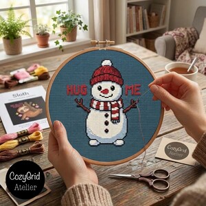 May include: A circular wooden embroidery hoop containing a cross-stitch pattern of a snowman wearing a red knit hat and a red and white striped scarf. The snowman is centered on a dark blue background with the red text HUG ME above it.