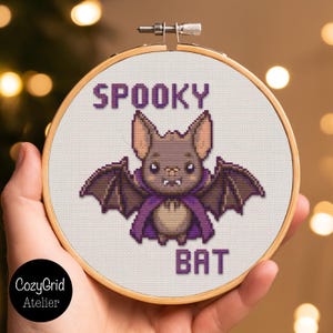 May include: A circular wooden embroidery hoop containing a cross-stitch pattern of a brown bat wearing a purple cape. The bat has large ears and small white fangs. The text SPOOKY is stitched in purple above the bat, and BAT is below it.