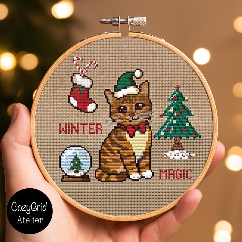 K&ouml;nnte beinhalten: Ein runder Holzstickrahmen mit einem Kreuzstichmotiv auf beigem Stoff. Das Design zeigt eine braune getigerte Katze mit gr&uuml;ner Weihnachtsm&uuml;tze und roter Fliege, einen roten Weihnachtsstrumpf, eine Schneekugel und einen Weihnachtsbaum mit dem Text WINTER MAGIC.