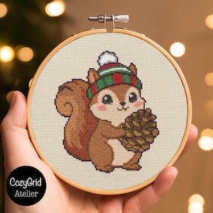 Op de afbeelding: Een kruissteekborduurwerk in een ronde houten borduurring met een bruine eekhoorn die een groen, rood en wit gestreepte wintermuts met pompon draagt. De eekhoorn houdt een bruine dennenappel vast op een crèmekleurige stoffen achtergrond.