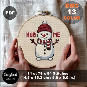 May include: A cross-stitch pattern in a wooden embroidery hoop featuring a snowman wearing a red hat and striped scarf with the text HUG ME. The design uses 13 DMC colors and measures 14.3 x 15.2 cm or 5.6 x 6.0 inches at 14 ct.