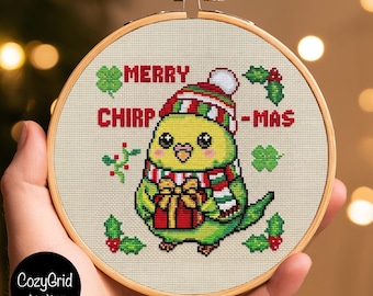 May include: A round wooden embroidery hoop containing a cross-stitch design of a green bird wearing a red, white, and green striped hat and scarf. The bird holds a red gift with a gold bow. Text reads MERRY CHIRP-MAS with holly accents.