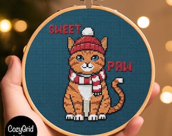 May include: A cross-stitch embroidery in a round wooden hoop featuring an orange tabby cat wearing a red knit hat and a red and white striped scarf. The dark blue background includes the red stitched text SWEET PAW.