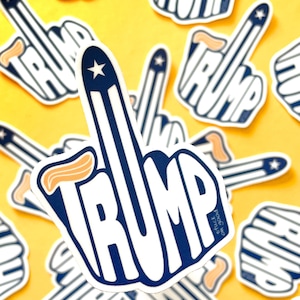 Puede incluir: Una pegatina blanca con un dedo medio azul y la palabra "TRUMP" escrita en letras azules. El dedo medio está decorado con un diseño de bandera estadounidense roja, blanca y azul. La pegatina está sobre un fondo amarillo.