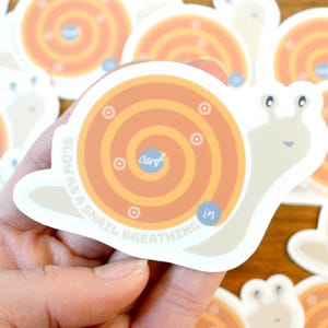 Puede incluir: Una pegatina de un caracol con una concha en espiral, la concha es naranja y amarilla con un c&iacute;rculo azul en el centro que dice "out". El caracol es beige y tiene una cara feliz. La pegatina dice "Slow as a snail breathing" alrededor del borde.