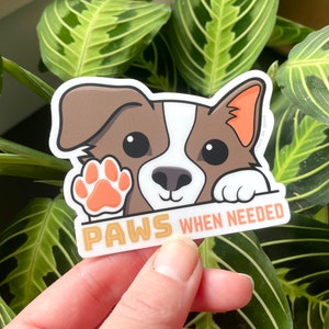 May include: A sticker featuring a cartoon dog with brown and white fur peeking over a white banner with the text "PAWS WHEN NEEDED" in orange and gold lettering. The dog has a brown nose, black eyes, and a pink paw print.