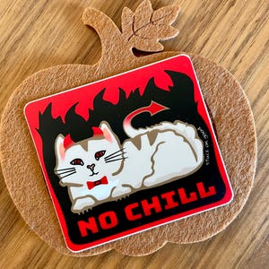Peut inclure: Un autocollant représentant un chat blanc avec des cornes de diable, un nœud papillon rouge et une queue de diable, sur un fond rouge et noir avec des flammes. Le texte "NO CHILL" est affiché en rouge. L'autocollant est placé sur une citrouille en feutre.