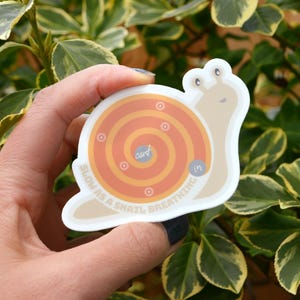 Puede incluir: Una pegatina de caracol blanca con una concha marr&oacute;n y un patr&oacute;n en espiral de color naranja y amarillo. El texto "Slow as a snail, breathing in" est&aacute; escrito alrededor de la espiral.