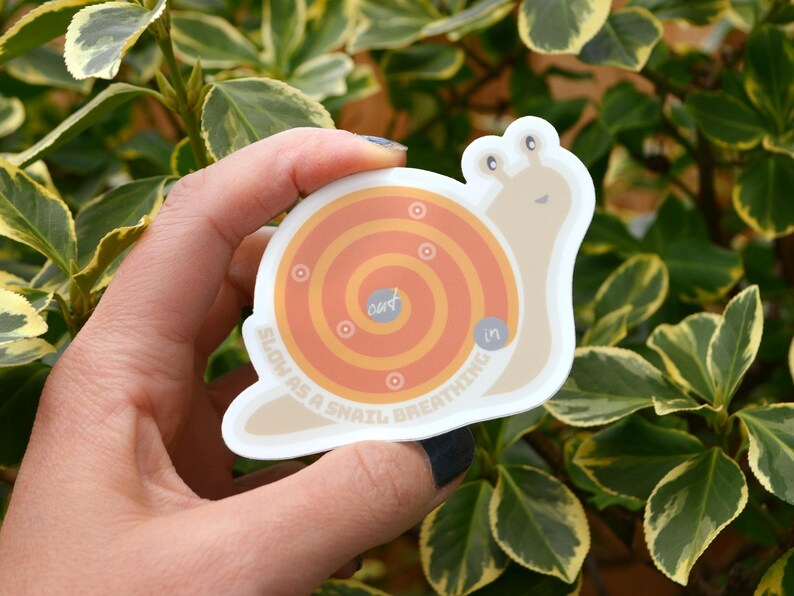 Puede incluir: Una pegatina de caracol blanca con una concha marr&oacute;n y un patr&oacute;n en espiral de color naranja y amarillo. El texto "Slow as a snail, breathing in" est&aacute; escrito alrededor de la espiral.