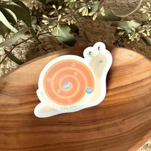 Puede incluir: Una pegatina blanca con un caracol de dibujos animados con una concha amarilla y rayas naranja y amarillas. El caracol lleva un objetivo con anillos naranja y amarillos y el texto "Respira hondo".