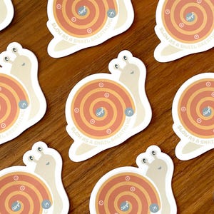 Puede incluir: Conjunto de nueve pegatinas en forma de caracol con un dise&ntilde;o en espiral de color naranja y amarillo. El texto "Slow as a snail, breathing in" est&aacute; impreso en cada pegatina.