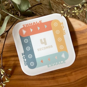 Puede incluir: Una tarjeta cuadrada blanca con un diseño colorido para ejercicios de respiración. La tarjeta tiene un diseño azul, verde y naranja con flechas y círculos. El texto en la tarjeta dice "INHALE", "HOLD", "EXHALE" y "4 SECONDS".
