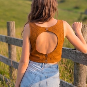 May include: A sleeveless, mustard-colored crop top featuring a textured fabric with small raised dots. The back of the garment has a large circular cutout, secured at the top and bottom with three brown buttons. The top is paired with light blue denim jeans.