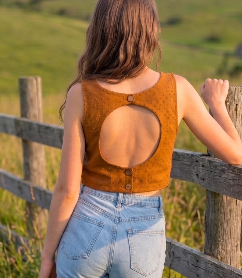 May include: A sleeveless, mustard-colored crop top featuring a textured fabric with small raised dots. The back of the garment has a large circular cutout, secured at the top and bottom with three brown buttons. The top is paired with light blue denim jeans.
