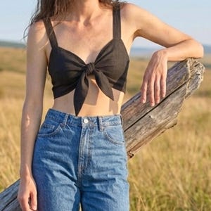 May include: A black cropped top featuring wide shoulder straps and a central front tie detail, paired with high-waisted blue denim jeans that have a classic button and zipper fly closure and visible stitching along the pockets and waistband.