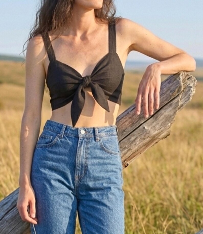May include: A black cropped top featuring wide shoulder straps and a central front tie detail, paired with high-waisted blue denim jeans that have a classic button and zipper fly closure and visible stitching along the pockets and waistband.