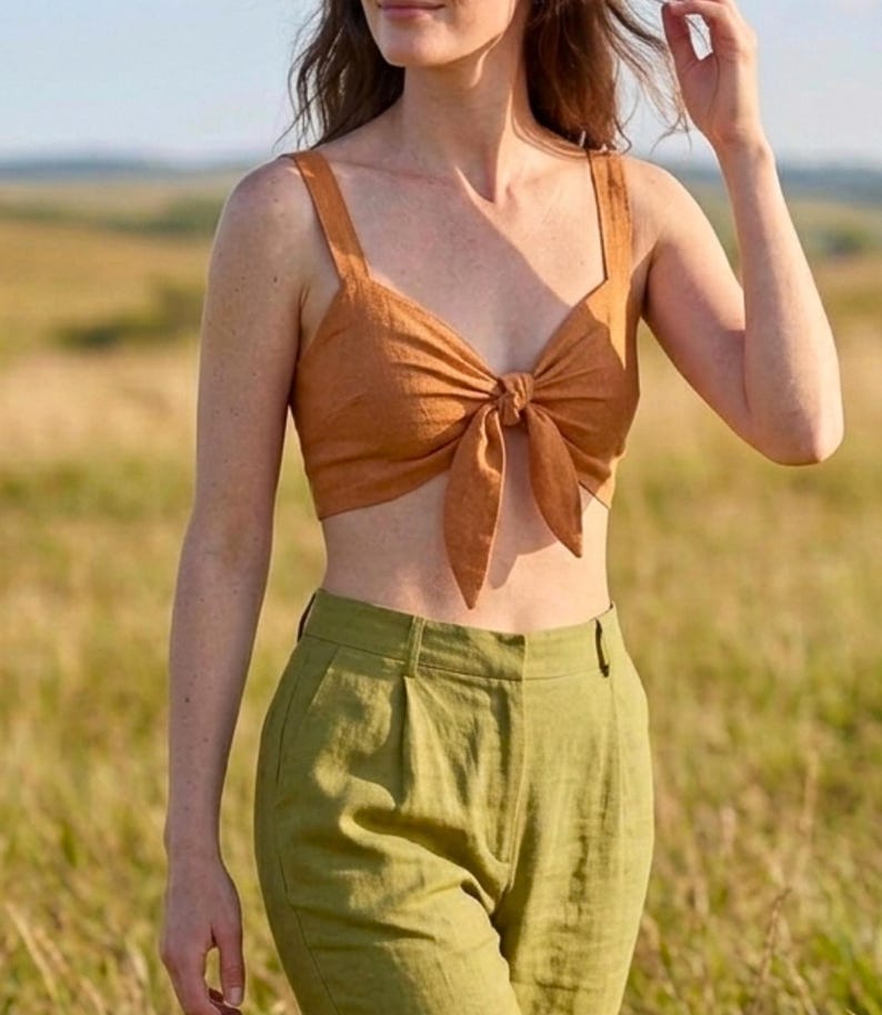 May include: A rust-colored cropped tank top featuring a front tie closure with wide shoulder straps, paired with high-waisted olive green trousers that have a pleated front design and a structured waistband with belt loops.