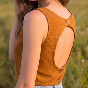 May include: A sleeveless mustard yellow top featuring a textured swiss dot fabric, a large circular open back cutout, and three round brown buttons positioned along the upper and lower back edges, paired with light blue denim jeans.