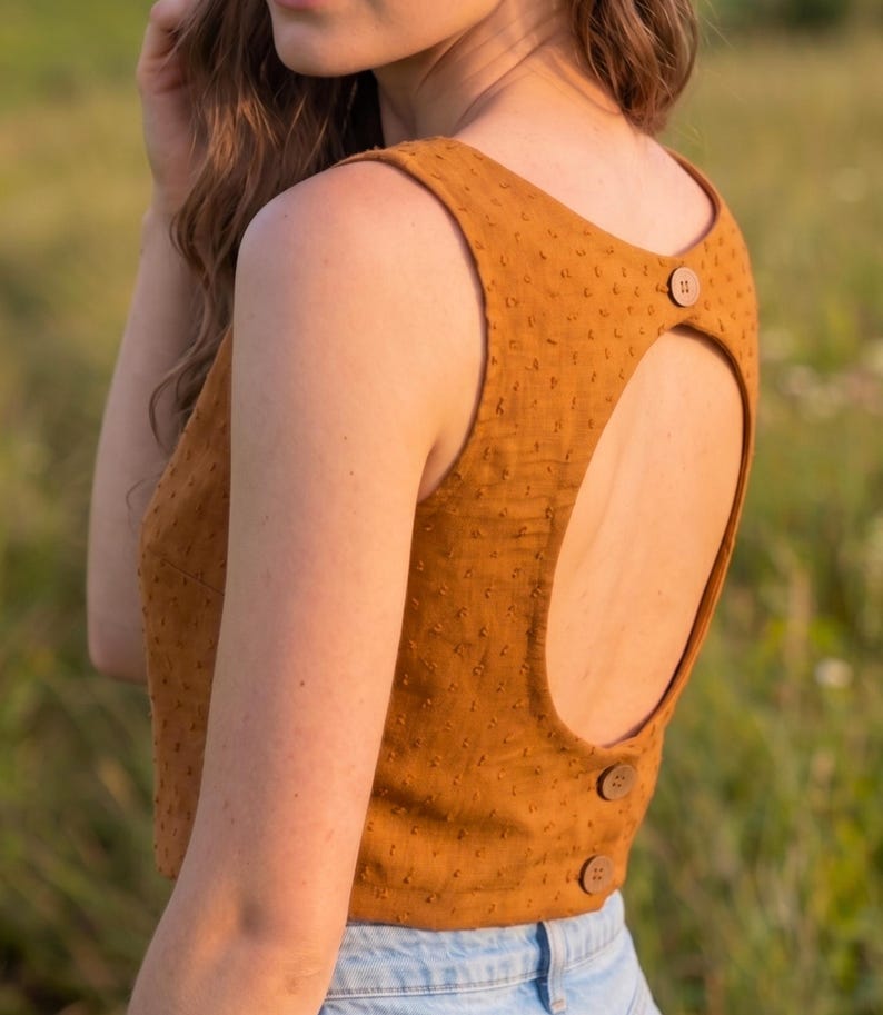 May include: A sleeveless mustard yellow top featuring a textured swiss dot fabric, a large circular open back cutout, and three round brown buttons positioned along the upper and lower back edges, paired with light blue denim jeans.