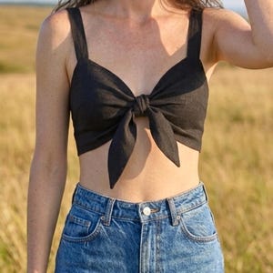 May include: A black cropped top featuring wide shoulder straps and a central front tie detail that creates a bow effect. The garment is paired with high-waisted blue denim jeans that have a classic button closure and visible metal rivets.