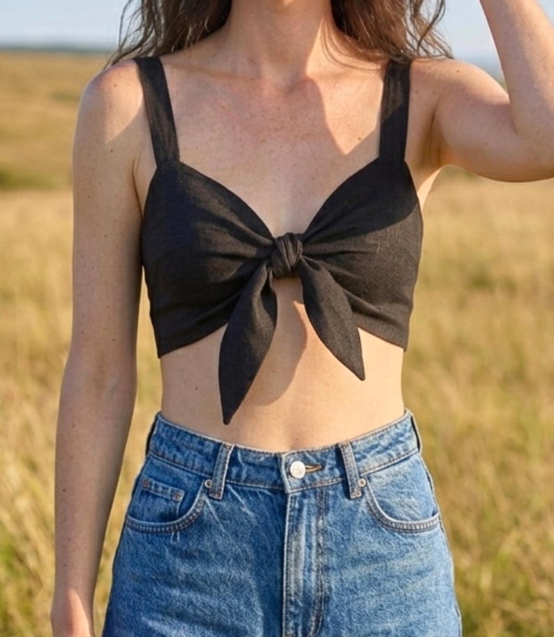 May include: A black cropped top featuring wide shoulder straps and a central front tie detail that creates a bow effect. The garment is paired with high-waisted blue denim jeans that have a classic button closure and visible metal rivets.