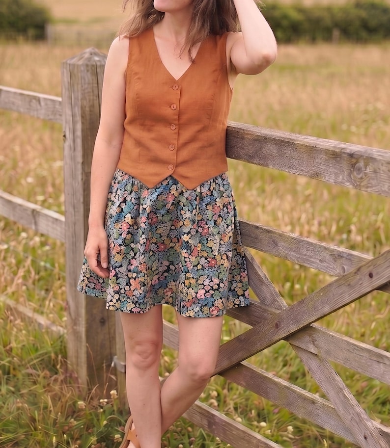 Pode incluir: Um colete sem mangas com decote em V numa cor laranja queimado s&oacute;lida com fecho de bot&otilde;es frontal, combinado com uma saia curta e evas&eacute; com um padr&atilde;o floral denso e multicolorido sobre um fundo preto com pequenas flores cor-de-rosa, azuis e amarelas.