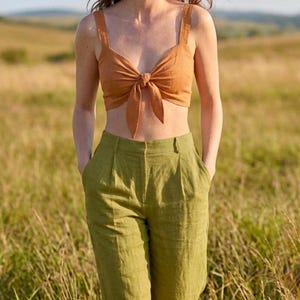 May include: A rust-colored cropped top featuring a front tie detail and wide shoulder straps, paired with high-waisted olive green trousers that have a pleated front design and side pockets. The outfit consists of a sleeveless top and long pants.