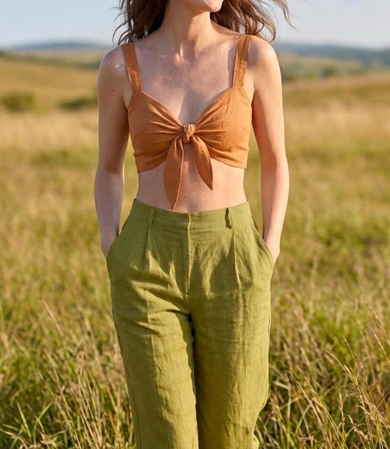 May include: A rust-colored cropped top featuring a front tie detail and wide shoulder straps, paired with high-waisted olive green trousers that have a pleated front design and side pockets. The outfit consists of a sleeveless top and long pants.