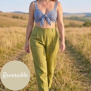 May include: A navy and white gingham print crop top with a front tie detail and thick straps, paired with high-waisted olive green trousers featuring a pleated front. A circular beige graphic overlay on the left side displays the text Reversible.