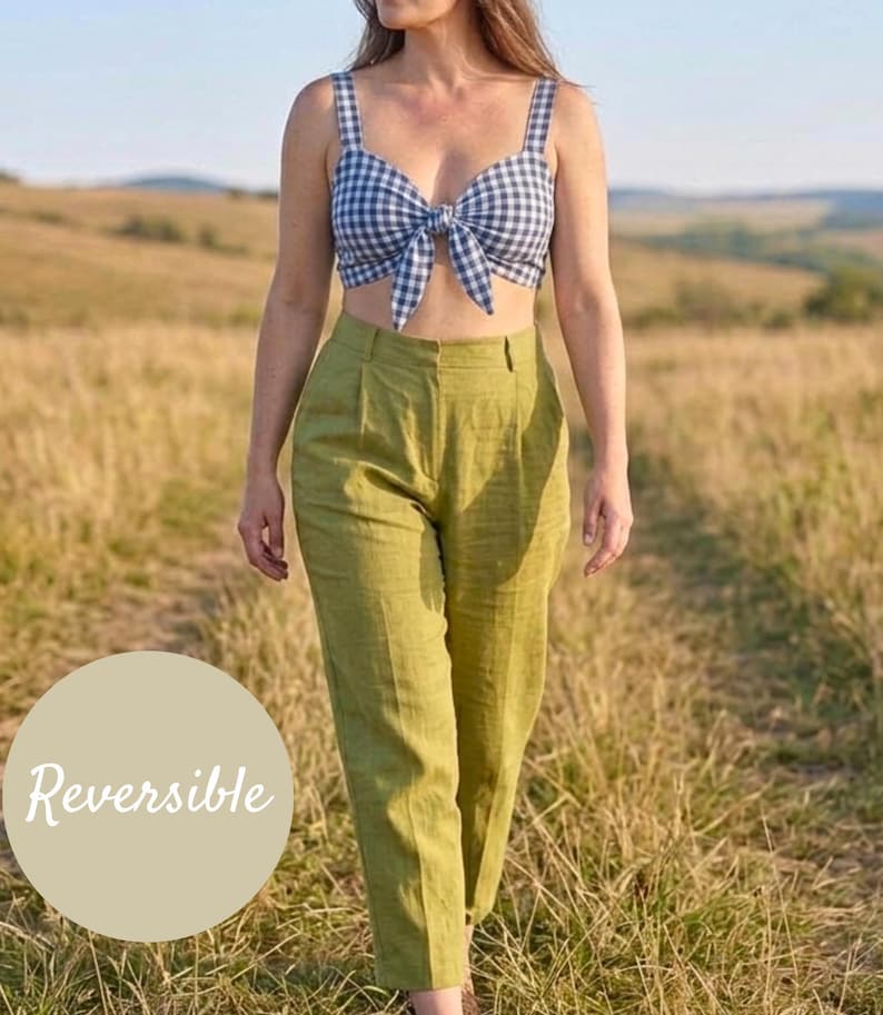 May include: A navy and white gingham print crop top with a front tie detail and thick straps, paired with high-waisted olive green trousers featuring a pleated front. A circular beige graphic overlay on the left side displays the text Reversible.