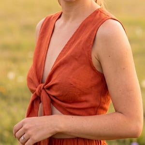May include: A sleeveless, rust-colored top featuring a deep V-neckline and a front tie closure at the waist. The garment is constructed from a textured fabric and is paired with matching high-waisted bottoms with an elasticized waistband.