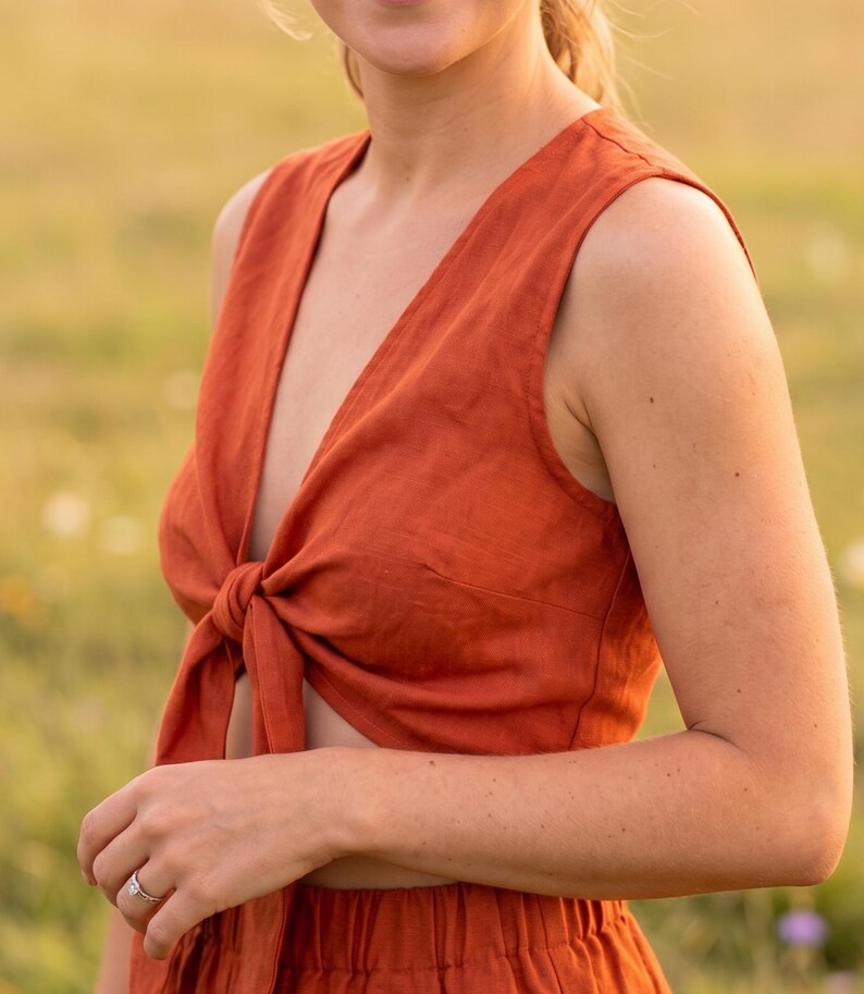 May include: A sleeveless, rust-colored top featuring a deep V-neckline and a front tie closure at the waist. The garment is constructed from a textured fabric and is paired with matching high-waisted bottoms with an elasticized waistband.