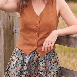 Pode incluir: Um colete sem mangas na cor ferrugem com decote em V e cinco bot&otilde;es a condizer na frente, combinado com uma saia com um padr&atilde;o floral denso e multicolorido sobre um fundo preto com tons de azul, rosa, amarelo e verde.