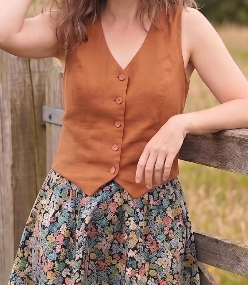 Pode incluir: Um colete sem mangas na cor ferrugem com decote em V e cinco bot&otilde;es a condizer na frente, combinado com uma saia com um padr&atilde;o floral denso e multicolorido sobre um fundo preto com tons de azul, rosa, amarelo e verde.