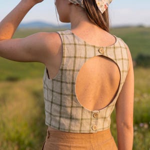 May include: A sleeveless beige crop top featuring a dark olive green grid pattern. The back of the garment has a large circular cutout, secured at the top and bottom with three small, round, light brown wooden buttons along the center seam.