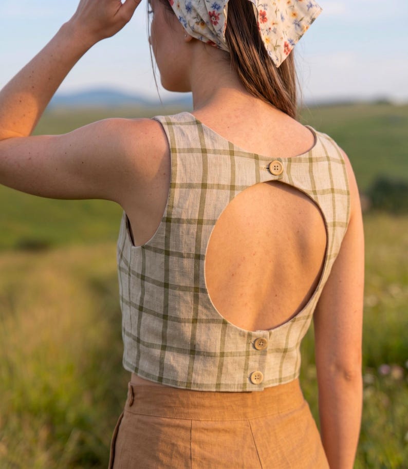 May include: A sleeveless beige crop top featuring a dark olive green grid pattern. The back of the garment has a large circular cutout, secured at the top and bottom with three small, round, light brown wooden buttons along the center seam.
