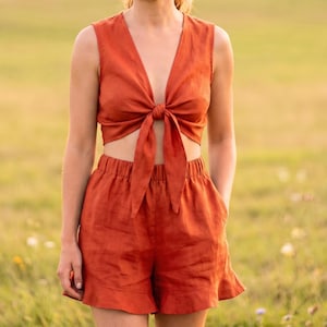May include: A two-piece set in a solid burnt orange color consisting of a sleeveless crop top with a deep V-neckline and a front tie closure, paired with matching high-waisted shorts featuring an elastic waistband and a ruffled hemline.