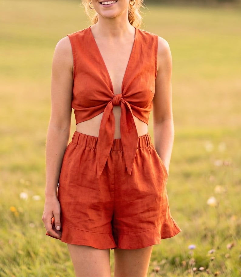 May include: A two-piece set in a solid burnt orange color consisting of a sleeveless crop top with a deep V-neckline and a front tie closure, paired with matching high-waisted shorts featuring an elastic waistband and a ruffled hemline.