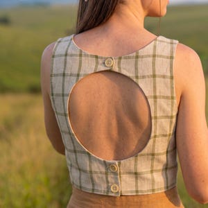 May include: A sleeveless beige crop top featuring a green plaid pattern, a large circular cutout on the back, and three small buttons along the center back closure. The garment is worn with a tan skirt and a floral patterned headscarf.