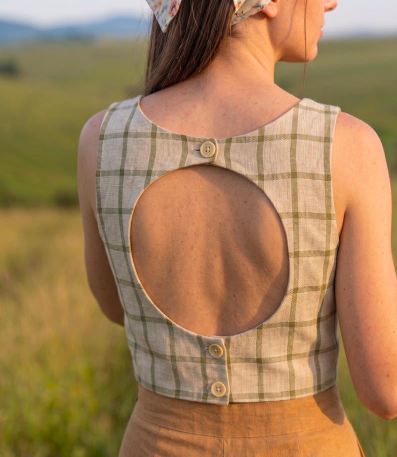 May include: A sleeveless beige crop top featuring a green plaid pattern, a large circular cutout on the back, and three small buttons along the center back closure. The garment is worn with a tan skirt and a floral patterned headscarf.