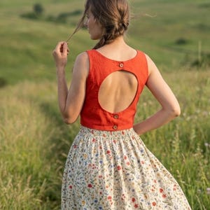 May include: A sleeveless, solid burnt orange crop top featuring a large circular back cutout, secured with three small brown buttons along the back closure, paired with a cream-colored skirt adorned with a delicate, multicolored floral print pattern.