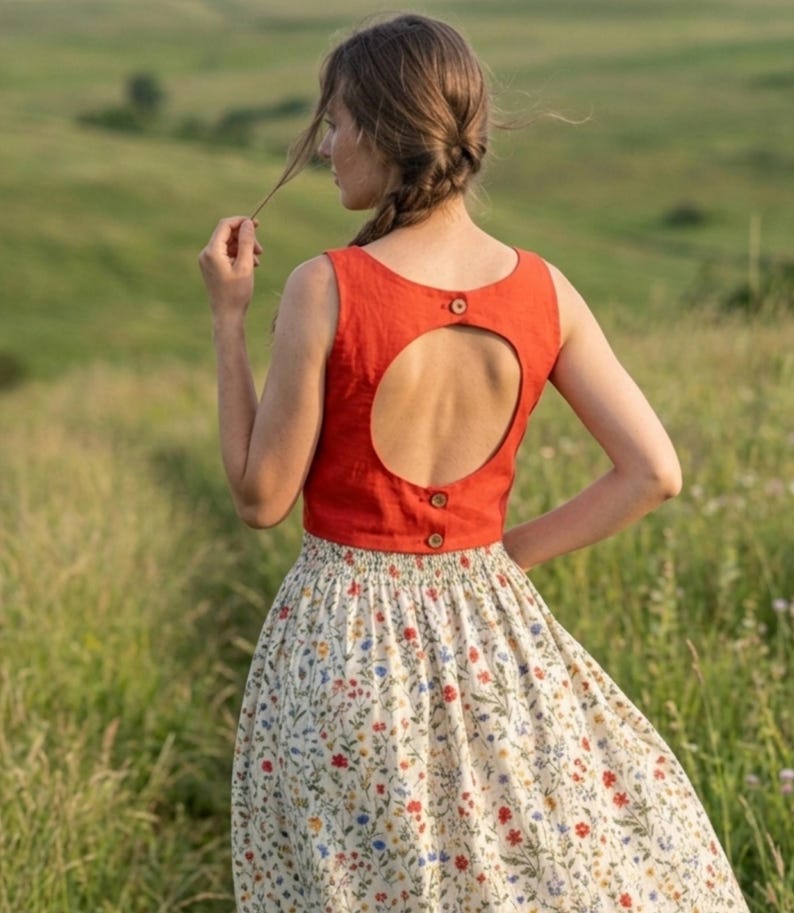 May include: A sleeveless, solid burnt orange crop top featuring a large circular back cutout, secured with three small brown buttons along the back closure, paired with a cream-colored skirt adorned with a delicate, multicolored floral print pattern.